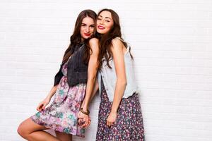 Close up   portrait of two pretty hipster  sisters  wearing boho colorful dress, stylish jeans jacket  and  baubles. Girls smile, have fun against  urban white wall. photo