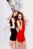 Two beautiful elegant women with red lips in evening black and red dress having fun. One keeping red stars balloons in her hand and smiling. Two woman at the party. Inside. White background. photo