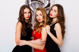 Three best friends celebrate  birthday indoor wearing elegant evening dress and have bright make up. Girls hugging and Showing signs with their hands. Looking at camera and smiling. Inside. photo
