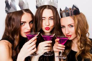 Three beautiful  elegant women celebrate hen-party and drinking  cocktails.  Best friends wearing black  evening dress ,crown  on head and clink glasses. Bright make up, red lips. Inside. photo