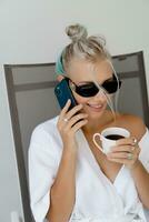 Close up portrait  of smiling  blond woman  in stylish sunglasses   and  white robe drinking cup of coffe in early morning. Resting in luxury hotel. photo