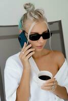 Close up portrait  of smiling  blond woman  in stylish sunglasses   and  white robe drinking cup of coffe in early morning. Resting in luxury hotel. photo