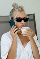 Close up portrait  of smiling  blond woman  in stylish sunglasses   and  white robe drinking cup of coffe in early morning. Resting in luxury hotel. photo