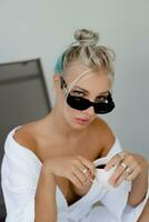 Close up portrait  of pleasure blond woman  in stylish sunglasses   and  white robe drinking cup of coffe in early morning. photo