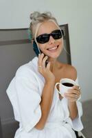 Close up portrait  of smiling  blond woman  in stylish sunglasses   and  white robe drinking cup of coffe in early morning. Resting in luxury hotel. photo