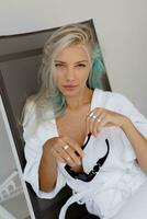 hermosa rubio mujer en blanco túnica Relajado en cubierta silla en lujo hotel. Perfecto piel, hermosa ojos. foto