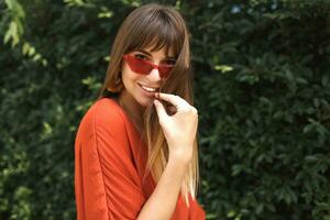 apareciendo morena mujer en elegante naranja atuendo y Paja sombrero Relajado en cubierta silla cerca piscina. fiesta y vacaciones concepto. foto