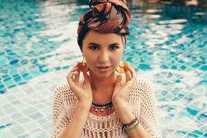 Woman with bright make up in bohemian outfit, turban, ethnic earrings, necklace and bracelets posing in swimming pool. photo