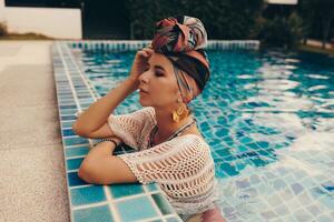 mujer con brillante hacer arriba en bohemio atuendo, turbante, étnico pendientes, collar y esposas posando en nadando piscina. foto