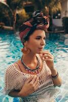 Woman with bright make up in bohemian outfit, turban, ethnic earrings, necklace and bracelets posing in swimming pool. photo