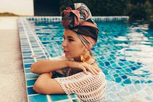 mujer con brillante hacer arriba en bohemio atuendo, turbante, étnico pendientes, collar y esposas posando en nadando piscina. foto