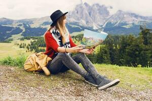 viajero joven mujer con mochila y sombrero sentado en césped y buscando Derecha dirección en mapa cerca increíble montañas y bosque vista. muestra un dedo a el cima. suave tonificado colores. foto