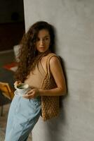 Lifestyle photo of pretty brunette woman in fashionable crop top and blue jeans holding cup of cofee. Modern interior.