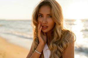 Close up portrait of beautiful blond woman in sexy swimwear posing on the beach in sunset light. Prtfect wavy hairs, tan skim body. Summer tropical mood. photo