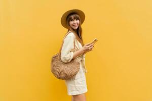 Smiling  carefree woman in straw hat and trendy summer linen  outfit using mobyle phone and posing on yellow background.  Vacation  and travel concept. photo