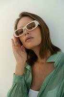 Close up portrait of stylish  woman in  sunglasses posing over wite background.  Casual outfit. Green linen blouse . white crop top and pants. photo