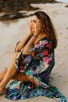 Geourgeous sensual  woman in pink swimsuit, and boho kimono  with tropical print  posing on  the beach at sunset. photo