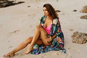 Geourgeous sensual  woman in pink swimsuit, and boho kimono  with tropical print  posing on  the beach at sunset. photo