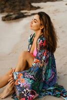 Geourgeous sensual  woman in pink swimsuit, and boho kimono  with tropical print  posing on  the beach at sunset. photo