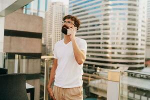 Handsome man with beard  talking by mobiyle phone while walking in big modern city with skyscrapers. Outdoors at sunset . Wearing white t shirt. photo