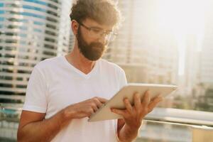 Handsome man in white t-shirt using  tablet , searching in internet, standing over moden city background with  skyscraper. photo