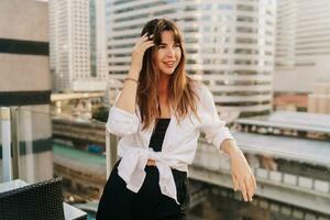 Pretty woman in casual outfit posing  on terrace in Bangkok. Modern buildings on background. photo