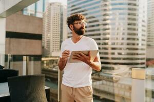 Stylish man with beard using tablet , standing on  roof top terrace in Bangkok. Modern buildings on background. photo