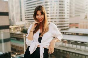 Pretty woman in casual outfit posing  on terrace in Bangkok. Modern buildings on background. photo