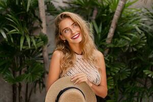 Beautiful blond woman in stylish knitted  boho dress posing in luxury tropical resort.  Perfect smile. Red lips. wavy hairs , straw hat. Vacation  mood. photo