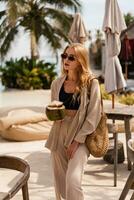 Pretty blond woman in stylish sunglasses dinking fresh coconut and posing in stylish beach club. Wearing summer linen costume. photo