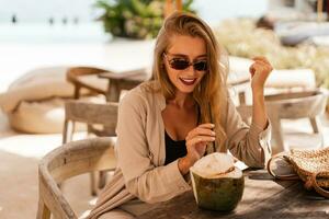 Stylish  blond woman in  sunglasses drinking fresh coconut and posing in stylish beach  cafe.Wearing summer linen costume. Travel and vacation mood. photo