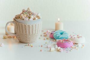 Cup of hot chocolate. Sprinkling chocolate on marshmallows in mug with cocoa. Colorful donuts on a white table surrounded with candles photo