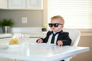 linda grave niñito chico vestido como un negocio hombre y en Gafas de sol se sienta en el cocina a el mesa para alimentación y murga para desayuno. pequeño jefe. ai generativo foto