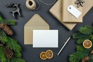 Christmas greeting card and envelope mockup with handmade gift box, pine cones and fir tree branches on dark background. Festive styled composition. Top view. photo