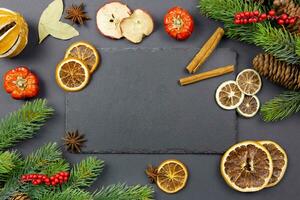 Winter dried fruits and culinary spice background mockup with christmas tree branches and pine cones on dark background with a black slate chalkboard for your text. Top view. Copy space. photo