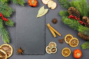 Winter dried fruits and culinary spice background mockup with christmas tree branches and pine cones on dark background with a black slate chalkboard for your text. Top view. Copy space. photo