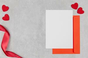 Blank greeting card mockup and red envelope on light concrete background with hearts confetti and ribbon. Valentines, mothers, womens day flat lay composition. Top view. Copy space. Love concept. photo