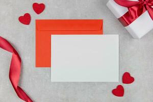 Blank greeting card mockup and red envelope on light concrete background with hearts confetti and ribbon. Valentines, mothers, womens day flat lay composition. Top view. Copy space. Love concept. photo