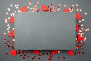 Red hearts confetti and black slate chalkboard mockup on dark background. Valentines, mothers, womens day, 8 march, birhday or wedding flat lay concept. Top view with copy space for your design. photo