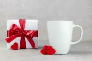 Classic white mug mockup with red hearts and gift box on light gray concrete background. Valentines, mothers, womens, 8 march or birthday concept. Side view. Copy space. photo