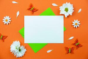 Blank greeting card mockup on colorful orange background with white daisy flowers and butterflies decoration. Spring flat lay composition. Top view. Copy space. photo