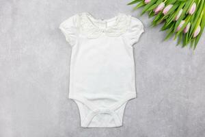 White baby girl bodysuit mockup flat lay with pink tulips flowers on the gray concrete background. Design onesie template, print presentation mock up. Top view. photo