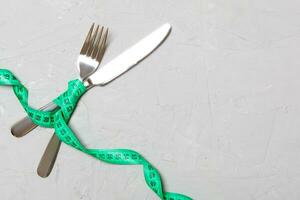 Top view of crossed knife and fork connected by measuring tape on gray cement background. Overeating and healthy diet concept photo