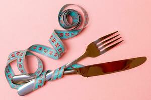 Composition of curled measuring tape, fork and knife on pink background with empty space for your design. Overweight concept photo
