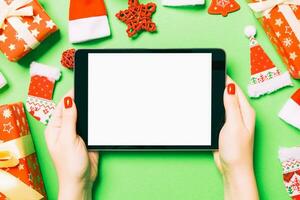 Top view of woman holding tablet in her hands on green background made of Christmas decorations. New Year holiday concept. Mockup photo
