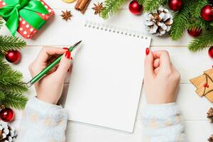 Top view of female hand writing in a notebook on wooden Christmas background. Fir tree and festive decorations. Wish list. New Year concept photo