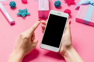 Top view of phone in female hand on festive pink background. Christmas decorations. New Year time holiday. Mockup photo