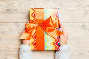 Woman hands give wrapped christmas or other holiday handmade present in colored paper . Present box, decoration of gift on yellow rustic wooden table, top view with copy space photo