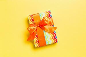 wrapped Christmas or other holiday handmade present in paper with orange ribbon on yellow background. Present box, decoration of gift on colored table, top view with copy space photo