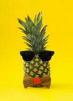 Creative tropical summer layout made of pineapple, sunglasses and wooden bow tie on light yellow background. Minimal summer fruit concept. Trendy exotic ananas idea. Pineapple aesthetic. photo
